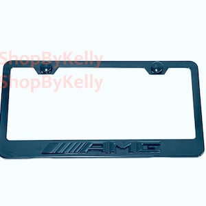 May include: A black rectangular license plate frame featuring the embossed AMG logo on the bottom edge. The frame includes two circular mounting points at the top and displays the text 3DShopByKelly in red across the upper portion of the frame.