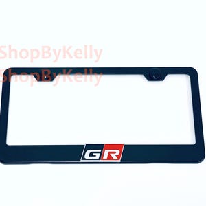 May include: A black rectangular license plate frame featuring the letters GR in white, set against a red and black background block centered at the bottom. The frame includes two mounting holes at the top and the text 3DShopByKelly.