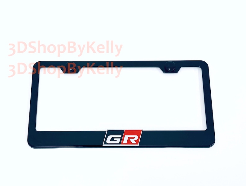 May include: A black rectangular license plate frame featuring the letters GR in white, set against a red and black background block centered at the bottom. The frame includes two mounting holes at the top and the text 3DShopByKelly.