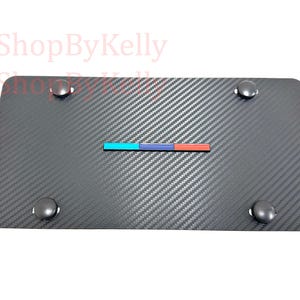May include: A rectangular license plate with a carbon fiber pattern. It features a horizontal stripe with blue, red, and teal colors. Four silver-colored fasteners are in each corner.