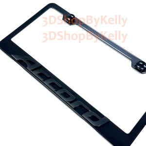 May include: A black license plate frame featuring the word ACCORD in raised, bold, black lettering along the bottom edge. The frame includes two black circular screw covers at the top and displays the text 3DShopByKelly in light orange.