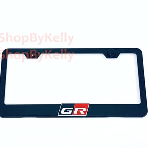 May include: A black rectangular license plate frame featuring the letters GR in white, set against a red and black background block at the bottom center. The frame includes two mounting holes at the top and the text 3DShopByKelly in light red.