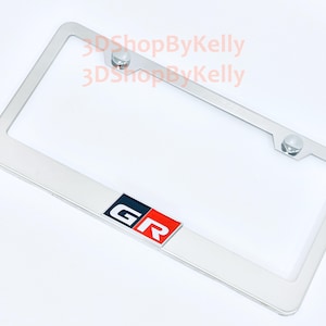 May include: A rectangular chrome license plate frame featuring a centered emblem with the letters GR in white, set against a black and red background. The frame includes two mounting holes at the top with silver hardware and the text 3DShopByKelly.