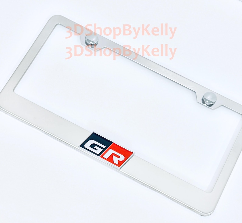 May include: A rectangular chrome license plate frame featuring a centered emblem with the letters GR in white, set against a black and red background. The frame includes two mounting holes at the top with silver hardware and the text 3DShopByKelly.