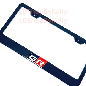 May include: A black rectangular license plate frame featuring a prominent GR logo at the bottom center. The logo consists of a white G and R on a red background. The frame includes two mounting holes at the top and the text 3DShopByKelly.