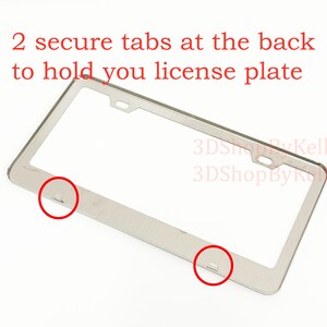 May include: A silver-colored metal license plate frame featuring two mounting holes at the top and two secure tabs at the bottom to hold a license plate. The text 2 secure tabs at the back to hold you license plate and 3DShopByKelly are visible.