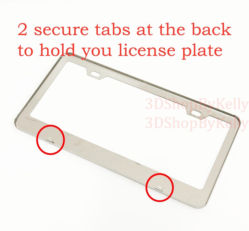 May include: A silver-colored metal license plate frame featuring two mounting holes at the top and two secure tabs at the bottom to hold a license plate. The text 2 secure tabs at the back to hold you license plate and 3DShopByKelly are visible.