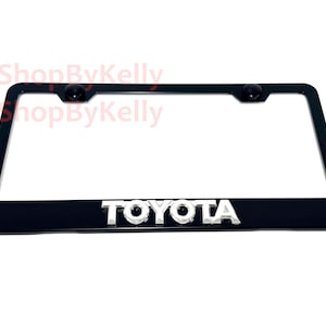 May include: Black license plate frame with the chrome "TOYOTA" logo. The frame has two black screw caps at the top. The text "3DShopByKelly" is visible in the upper left and upper right corners.