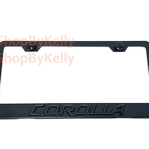 May include: A black rectangular license plate frame featuring the word COROLLA embossed in capital letters along the bottom edge. The frame includes two circular black mounting caps positioned at the top corners for secure vehicle attachment.