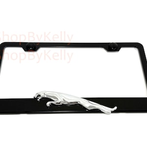 May include: Black license plate frame with a chrome Jaguar emblem. The frame has a rectangular shape with rounded corners and two black screw caps at the top. The Jaguar emblem is a silver-colored leaping cat.