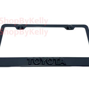 May include: Black license plate frame with the embossed word "TOYOTA" at the bottom. The frame has a rectangular shape with rounded corners and two black screw covers at the top.