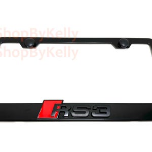 May include: Black license plate frame with the letters "RS3" in black and red. The frame has two black fasteners at the top. The frame is rectangular and has rounded corners.