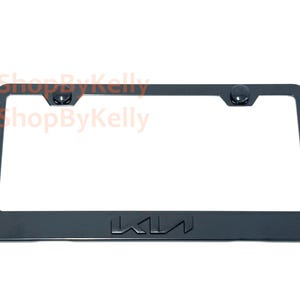 May include: A rectangular black license plate frame featuring the Kia logo embossed in the center of the bottom edge. The frame includes two circular black screw covers positioned at the top corners. The text 3DShopByKelly is visible in orange.