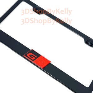 May include: A black rectangular license plate frame featuring a prominent red and black GR emblem centered at the bottom. The frame includes two black circular screw covers at the top. The text 3DShopByKelly is visible in light orange.