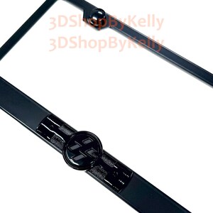 May include: A black license plate frame featuring a circular emblem with a stylized black logo in the center. The frame has a smooth finish with a raised circular detail at the top and a textured rectangular section surrounding the central emblem.
