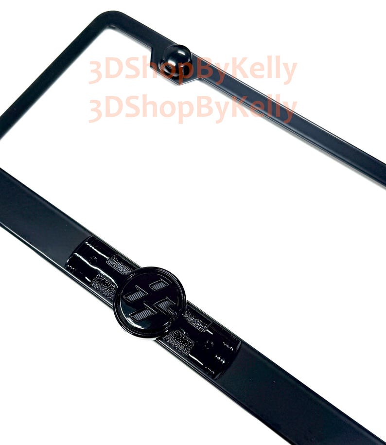 May include: A black license plate frame featuring a circular emblem with a stylized black logo in the center. The frame has a smooth finish with a raised circular detail at the top and a textured rectangular section surrounding the central emblem.