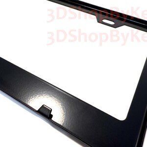 May include: A glossy black license plate frame with a rectangular shape and rounded corners. The frame features a centered mounting hole at the top and a small tab at the bottom. The text 3DShopByKelly is printed in red across the upper section.