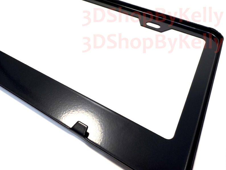 May include: A glossy black license plate frame with a rectangular shape and rounded corners. The frame features a centered mounting hole at the top and a small tab at the bottom. The text 3DShopByKelly is printed in red across the upper section.