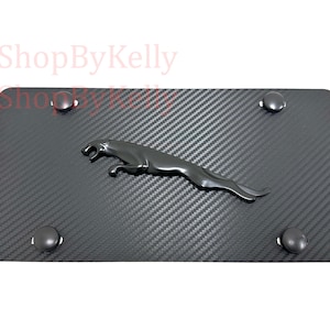 May include: A black license plate with a carbon fiber pattern and a dark gray Jaguar emblem. The plate has four dark gray mounting hardware pieces. The background is white.