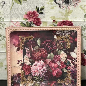 May include: A rectangular decorative tray with a pink beaded frame. The tray's interior features a floral design with pink, burgundy, and cream-colored flowers. Gold filigree accents adorn the corners, adding an elegant touch.