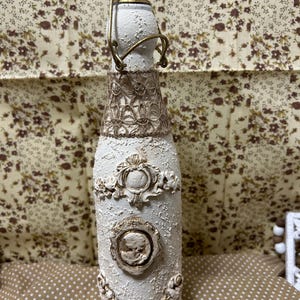 May include: A decorative white bottle with a textured surface and ornate embellishments. The bottle features a gold-colored metal closure and is adorned with a brown lace-like band and framed relief details, including a skull. The bottle is approximately 10 inches tall.