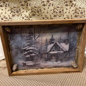 May include: Wooden framed winter scene featuring a snow-covered cottage and trees. The frame is made of light brown wood and has decorative pine cones on the corners. The artwork is a print of a winter landscape.
