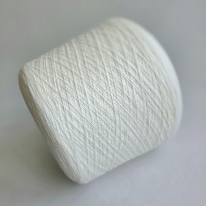 May include: A cylindrical spool of off-white yarn featuring a distinct cross-hatched winding pattern. The thread is tightly wound around the core, creating a textured surface with visible diagonal lines throughout the entire structure of the spool.