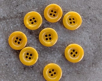 May include: Seven round, yellow buttons with four holes each are arranged on a gray surface. The buttons have a glossy finish and a slightly translucent appearance, suggesting they are made of plastic or resin.