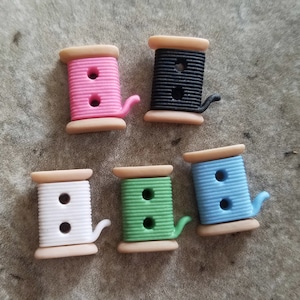 May include: Six colorful plastic buttons shaped like spools of thread. The buttons are pink, black, white, green, and blue. Each button has two holes.