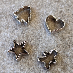 May include: Four silver metal cookie cutters in the shapes of a Christmas tree, a heart, a star, and a gingerbread person.