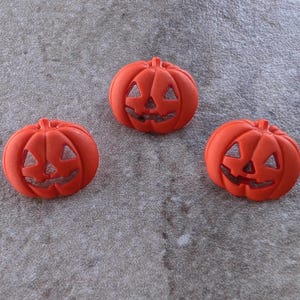 May include: Three bright orange plastic buttons shaped like jack-o'-lanterns. Each button features a carved face with triangular eyes, a triangular nose, and a wide, jagged mouth. The surface of each pumpkin button is smooth and matte.