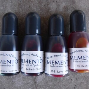 Puede incluir: Cuatro botellas peque&ntilde;as de tinta de tinte resistente a la decoloraci&oacute;n Memento. Las botellas tienen tapas de color gris oscuro y etiquetas blancas con texto negro. Los colores de tinta incluyen tonos de verde, rojo y naranja. Las etiquetas dicen "Water-Based, Acid Free".
