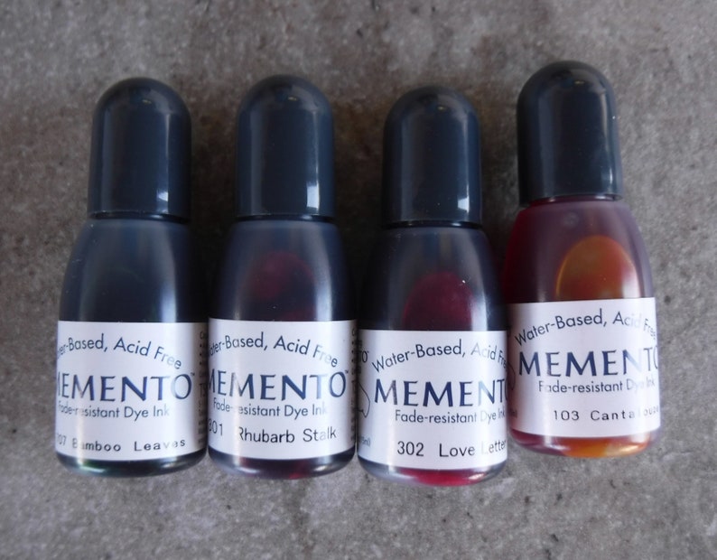 Puede incluir: Cuatro botellas peque&ntilde;as de tinta de tinte resistente a la decoloraci&oacute;n Memento. Las botellas tienen tapas de color gris oscuro y etiquetas blancas con texto negro. Los colores de tinta incluyen tonos de verde, rojo y naranja. Las etiquetas dicen "Water-Based, Acid Free".
