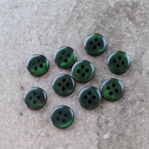 May include: A collection of ten round, dark green buttons, each featuring a smooth, slightly glossy finish and four centered holes for sewing. The buttons are arranged in a scattered pattern, displaying a consistent circular shape and deep forest green hue.