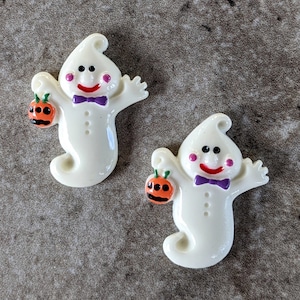 May include: Two white ghost figurines with black eyes, pink cheeks, and purple bow ties. Each ghost is holding a small orange pumpkin.