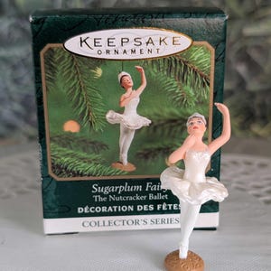 May include: A Hallmark Keepsake Ornament box featuring a Sugarplum Fairy from The Nutcracker Ballet, accompanied by a small figurine of a ballerina in a white tutu and slippers standing on a round brown base with one arm raised above the head.