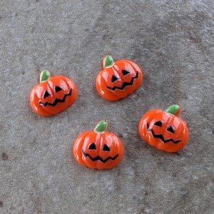 May include: Four small, glossy orange pumpkin-shaped charms, each featuring a carved jack-o'-lantern face with black triangular eyes and a jagged mouth. Each pumpkin has a small, smooth green stem at the top, providing a vibrant contrast.