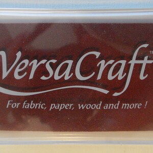 May include: A clear plastic container with a brown ink pad inside. The ink pad has the text "VersaCraft&trade;" in white lettering and the words "For fabric, paper, wood and more!" in smaller white lettering.