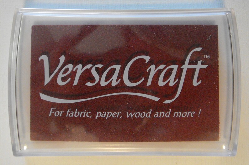 May include: A clear plastic container with a brown ink pad inside. The ink pad has the text "VersaCraft&trade;" in white lettering and the words "For fabric, paper, wood and more!" in smaller white lettering.