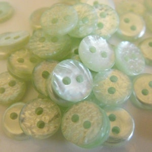 May include: A collection of pale green, round buttons with two or three holes. The buttons have a pearlescent sheen and a slightly textured surface. These small, decorative buttons are suitable for sewing and craft projects.