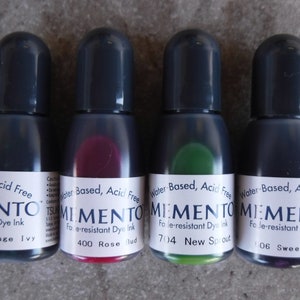Puede incluir: Cuatro botellas peque&ntilde;as de tinta de tinte Memento a base de agua, sin &aacute;cido y resistente a la decoloraci&oacute;n. Las botellas tienen tapas de color gris oscuro y contienen tintas en tonos azul, rojo, verde y morado. Cada botella tiene una etiqueta blanca.