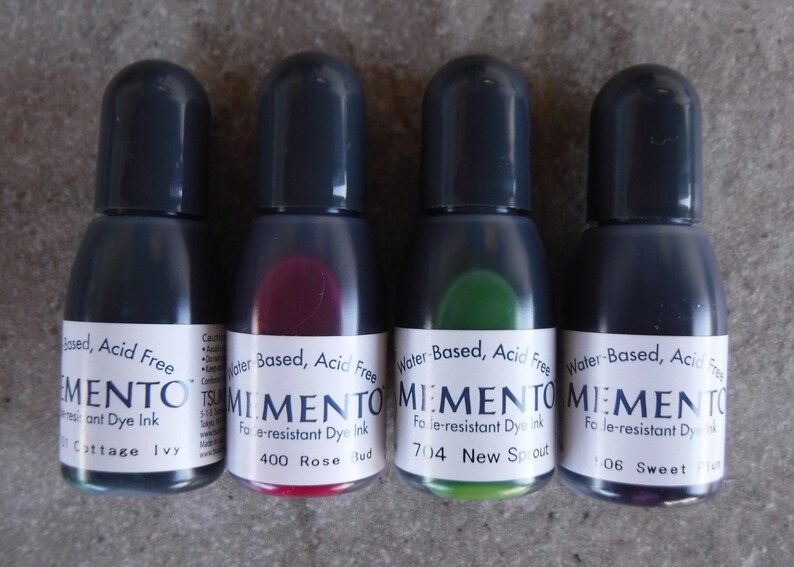 Puede incluir: Cuatro botellas peque&ntilde;as de tinta de tinte Memento a base de agua, sin &aacute;cido y resistente a la decoloraci&oacute;n. Las botellas tienen tapas de color gris oscuro y contienen tintas en tonos azul, rojo, verde y morado. Cada botella tiene una etiqueta blanca.