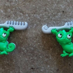 May include: Two green chameleon figurines holding up white combs. The chameleons have large eyes and are holding the combs with their front legs.