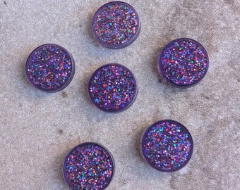 May include: Five circular flat buttons featuring a deep purple base filled with multi-colored iridescent glitter particles. Each button has a smooth, glossy finish and a consistent round shape, arranged in a scattered pattern across the frame.