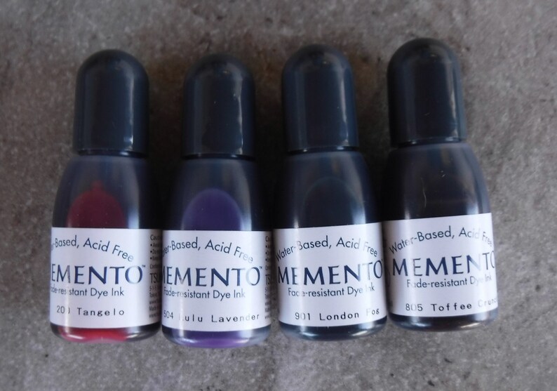 Puede incluir: Cuatro peque&ntilde;os frascos de tinta con tapas negras y etiquetas blancas. Los frascos contienen tinta roja, morada, azul y marr&oacute;n. Las etiquetas dicen "MEMENTO" y "Fade-resistant Dye Ink". Los frascos de tinta est&aacute;n etiquetados con los colores Tangelo, Lulu Lavender, London Fog y Toffee Crunch.