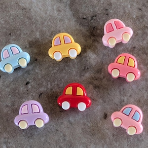 May include: A collection of seven colorful toy car embellishments. The cars are in various colors, including blue, yellow, pink, red, and purple, with white wheels and contrasting window details. The cars are arranged on a gray surface.