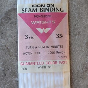 May include: A package of white iron-on seam binding by Wrights. The label features a pink triangle with the text IRON ON SEAM BINDING, NON-SHRINK, WRIGHTS, 3 Yds., 35 cents, TURN A HEM IN MINUTES, WOVEN EDGE, 100% RAYON, GUARANTEED COLOR FAST, 608, and WHITE 30.