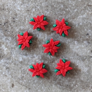 May include: Six red poinsettia flower embellishments with green leaves are arranged on a textured, neutral-toned surface. The flowers have detailed petals and are likely for crafting or holiday decor.