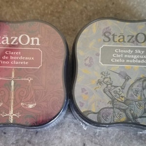 May include: Two ink pads with the brand name "StazOn" printed on the front. The ink pad on the left is red and says "Claret Vin de bordeaux Vino clarete". The ink pad on the right is blue and says "Cloudy Sky Ciel nuageux Cielo nublado".