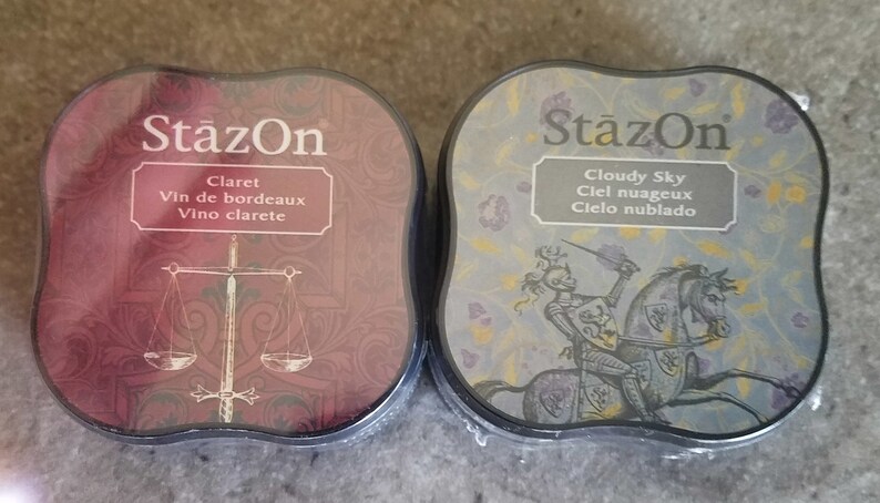 May include: Two ink pads with the brand name "StazOn" printed on the front. The ink pad on the left is red and says "Claret Vin de bordeaux Vino clarete". The ink pad on the right is blue and says "Cloudy Sky Ciel nuageux Cielo nublado".